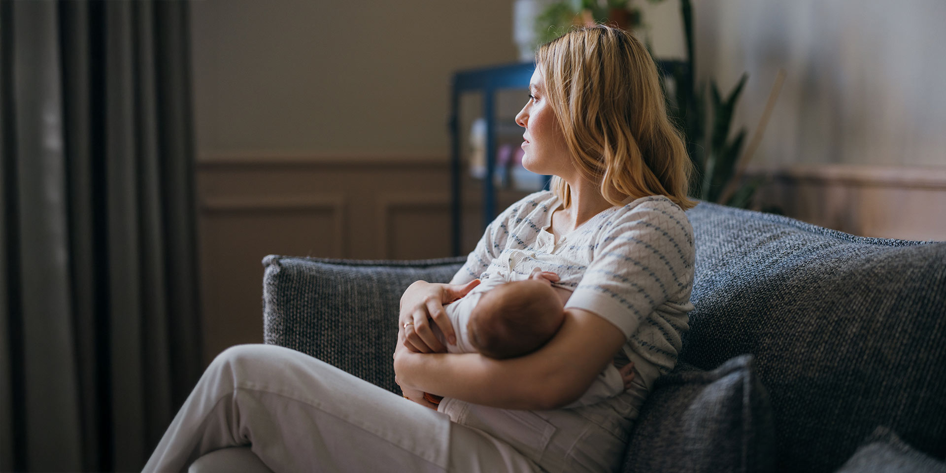 eine nachdenkliche mutter füttert ihr süsses baby, während sie es hält. sie sitzt auf dem gemütlichen sofa zu hause. symbolbild postpartale depression