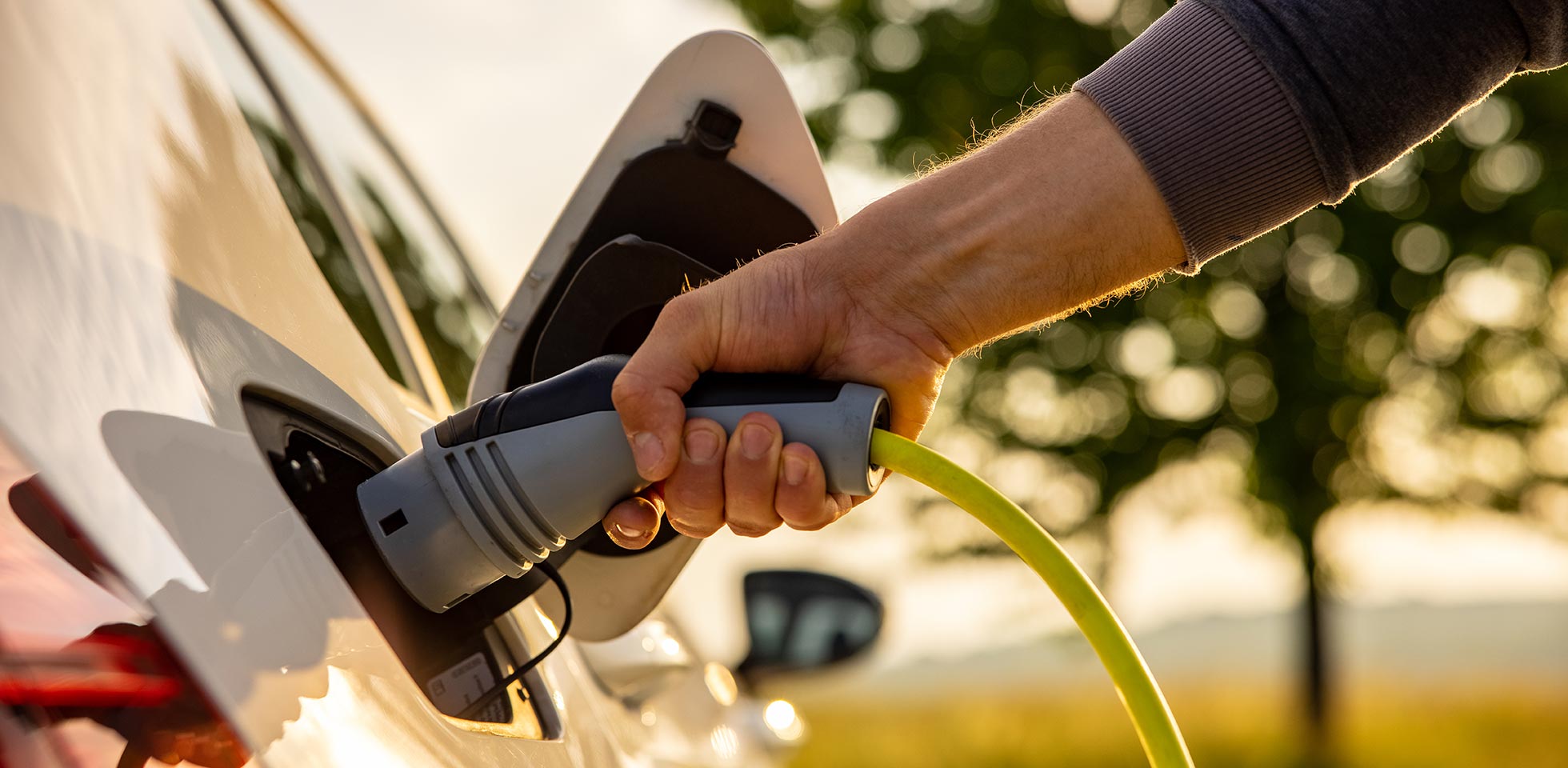 hand eines mannes,  ein stromkabel in ein elektroauto einführt, um ein umweltfreliches fahrzeug in einer grünen landschaft aufzuladen
