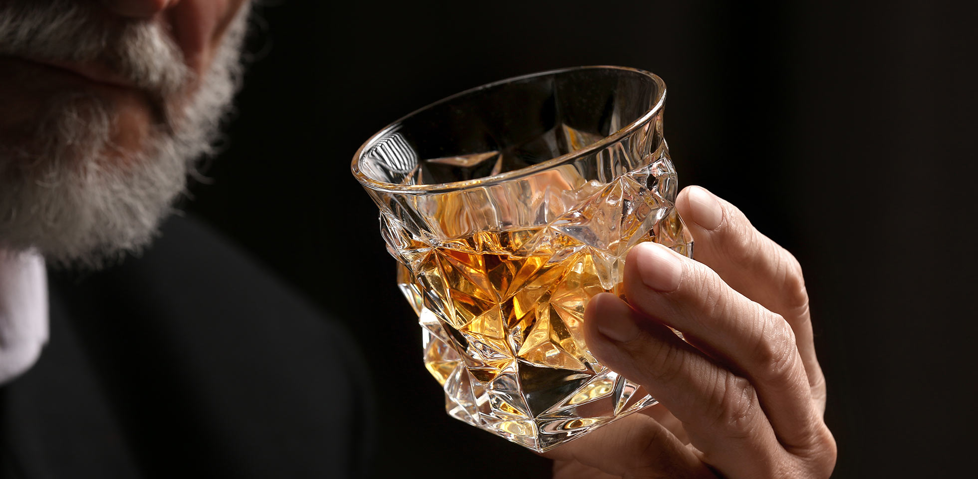 mann hält ein glas whiskey  eiswürfeln vor einem dunklen hintergr, nahaufnahme. symbolbild lebensgenuss