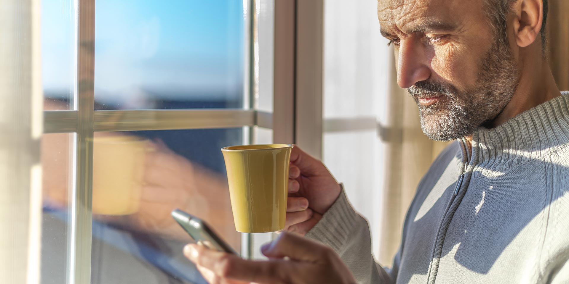 close-up shot of smiling adult man's face with beard holding a cup in one hand and a mobile phone - which he is looking at - in the other hand next to window with morning light, regulatorik