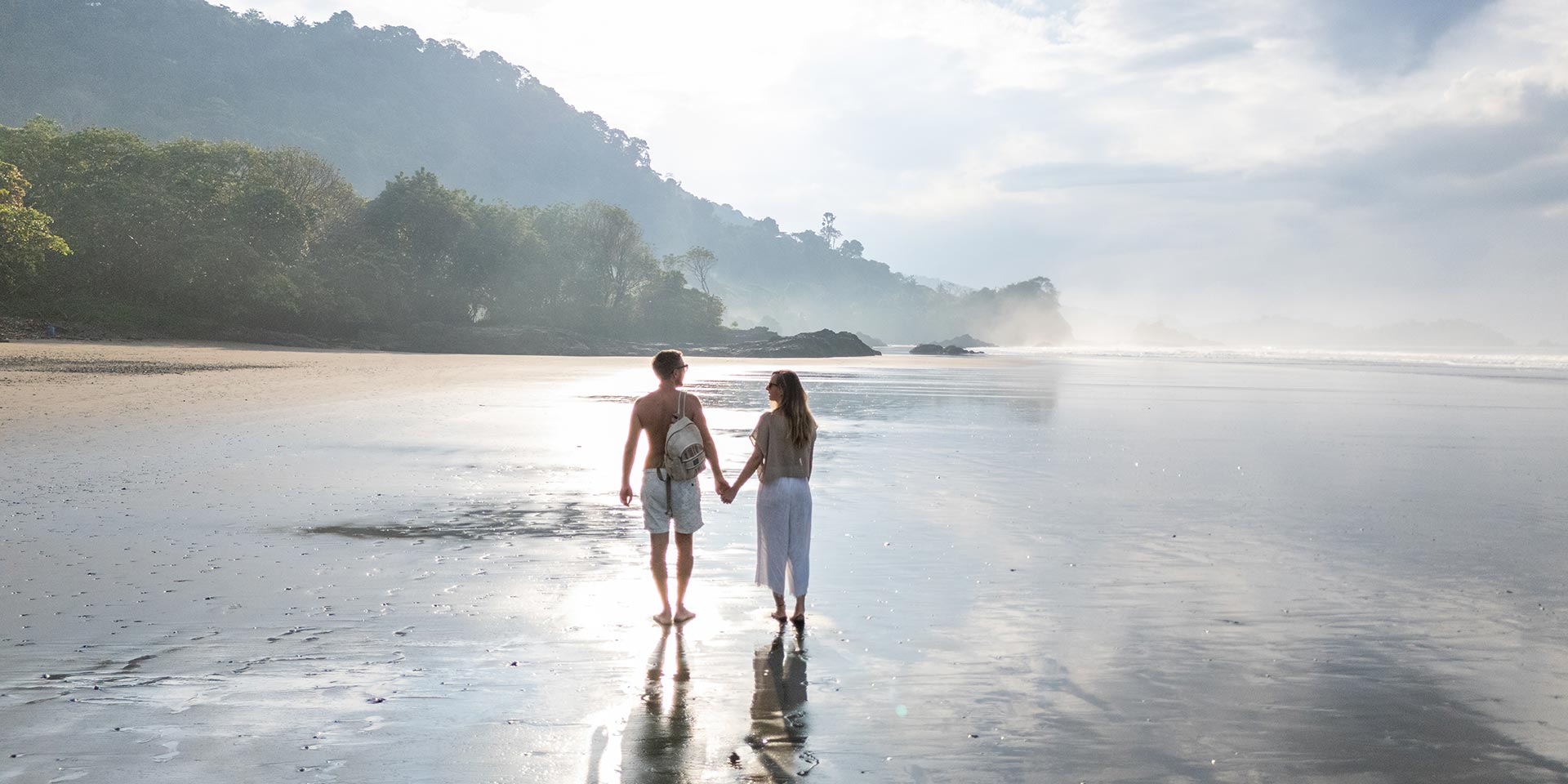 couple strolling on a tranquil beach at sunrise. silhouettes reflected in wet sand by the ocean. serene coastal landscape with misty horizon and lush greenery.